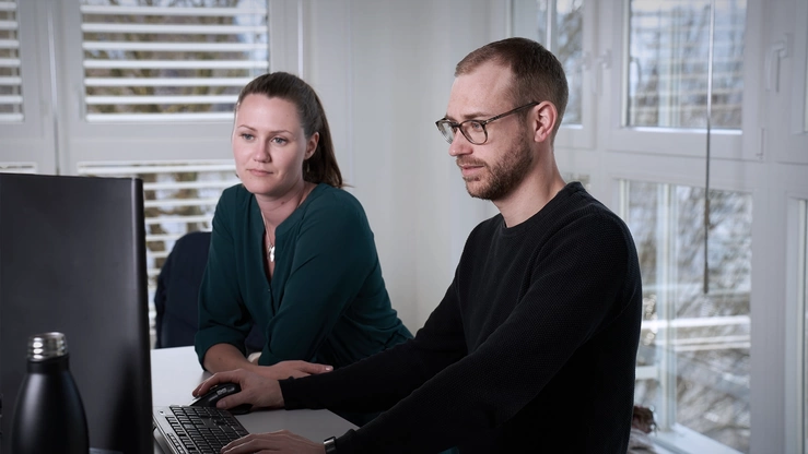 Zwei Mitarbeitende hinter einem PC-Screen